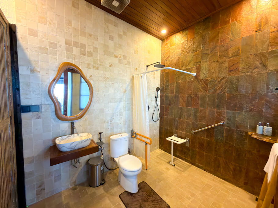 A spacious walk-in bathroom combines cream colored stone tile walls and floor with a feature wall of earthy rust and brown slate tiles under a warm wooden ceiling.