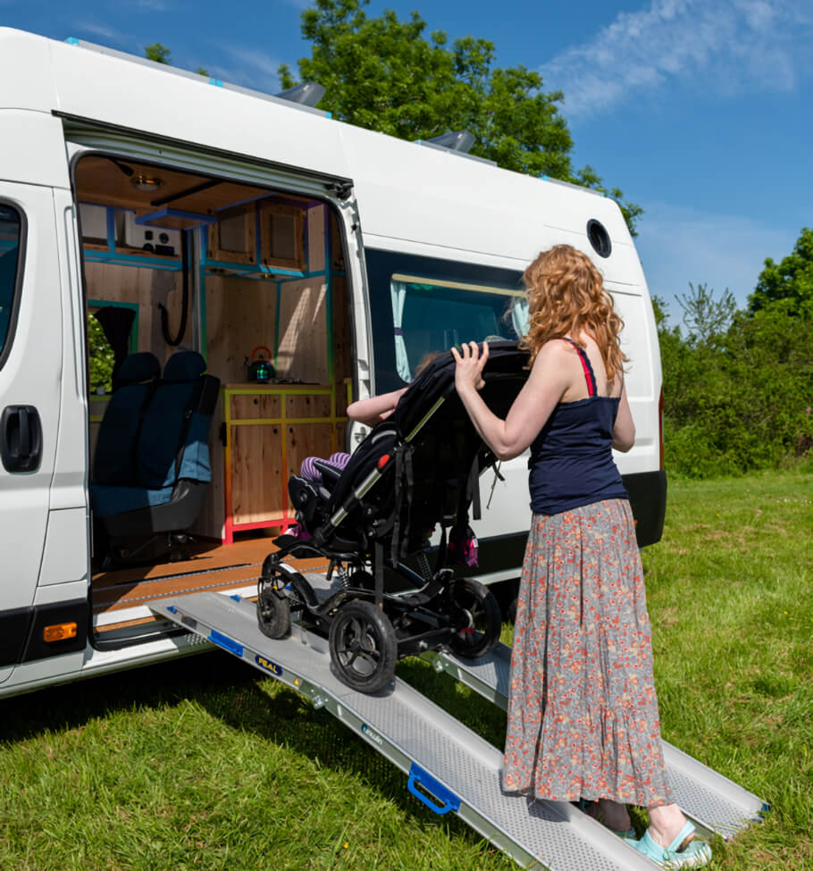 The image shows a woman with curly red hair pushing a stroller with a child up a metal ramp into a white camper van parked on a grassy field. The van's side door is open, revealing a cozy, wood-paneled interior with seats and shelves. The weather is sunny, with a bright blue sky and green trees in the background.