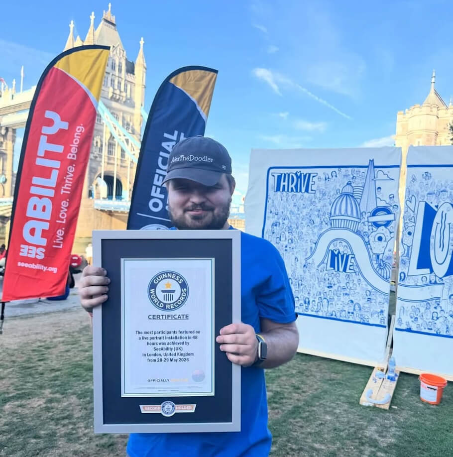 This image shows Alex the Doodler standing outdoors in front of London’s iconic Tower Bridge, proudly holding a framed Guinness World Records certificate awarded for a project supporting people with visual impairments.