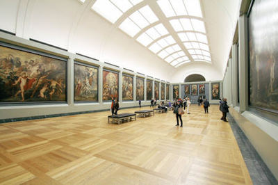 Photo of the interior of an art exhibition gallery showing people looking at paintings hanging on the walls under a domed shape roof.