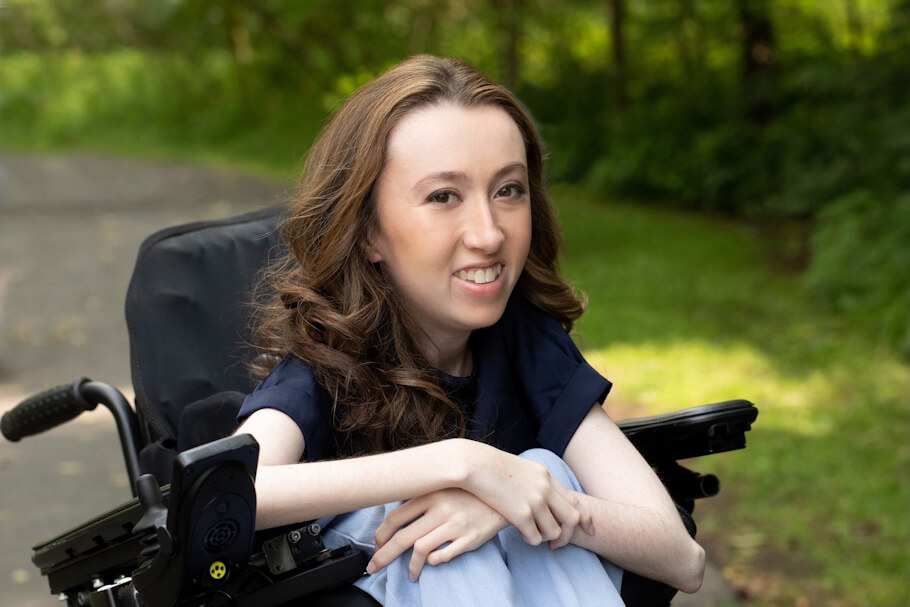Avery Roberts sits in a powered wheelchair on a paved path surrounded by lush green trees and foliage, smiling warmly toward the camera.