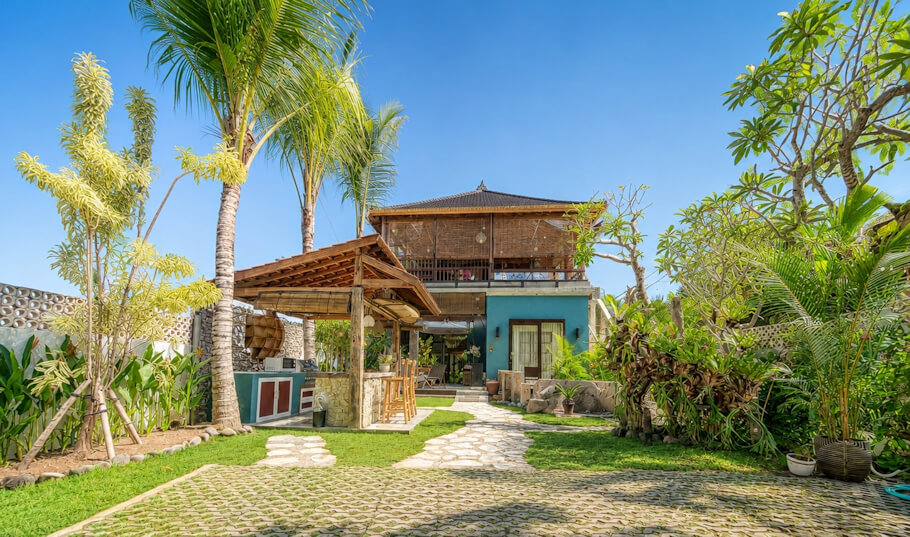 Villa exterior and garden: Under a bright blue sky, a two story villa with a traditional Balinese hip roof and woven bamboo upper walls rises behind a lush tropical garden. Tall palm trees and flowering frangipani frame the scene, and a flat stone pathway crosses a neatly kept lawn toward the house.