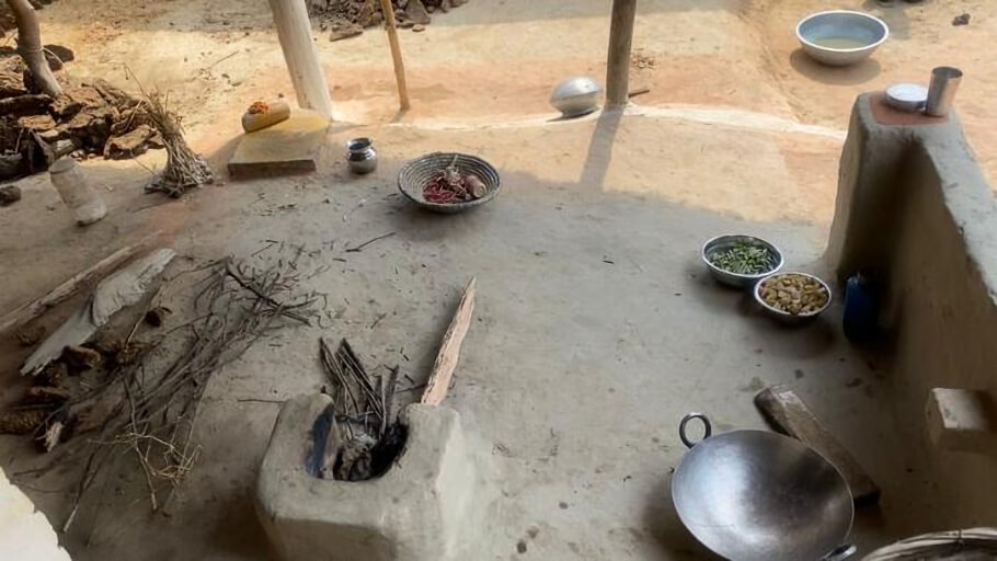 This image shows a simple traditional outdoor cooking area in a rural home in Uttar Pradesh. In the foreground is a clay-and-mud wood-fired stove built low to the ground, with pieces of firewood inserted into its opening for cooking.