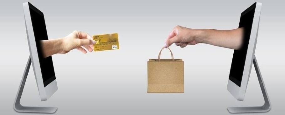 Two computers face each other, a hand holding a credit card extends from one screen towards the other screen where another hand holding a brown shopping bag reaches out in exchange.