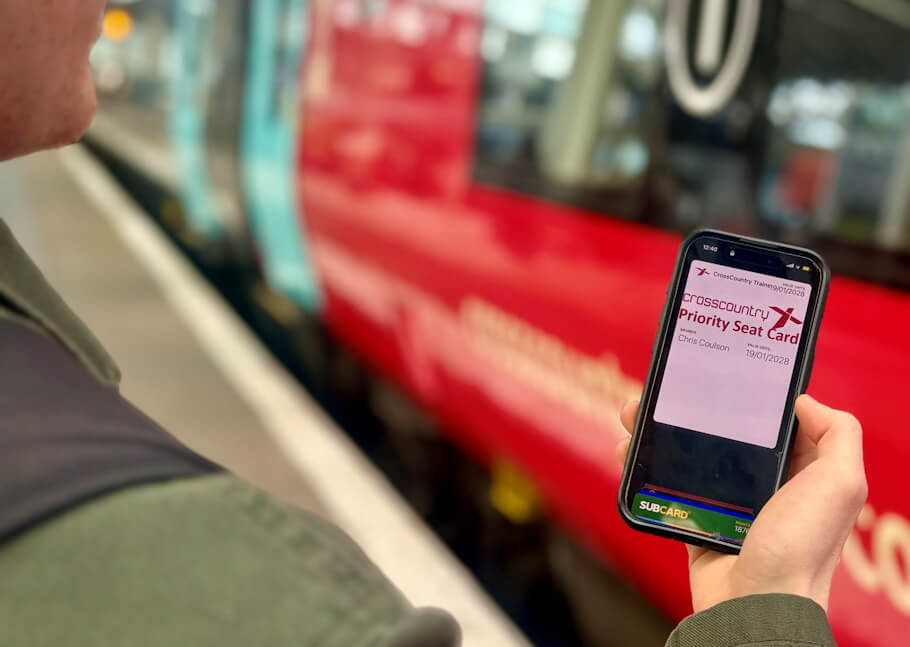 A person seen from over their shoulder, wearing a green jacket, stands on a train station platform holding a smartphone in their right hand and looking down at its screen, while a red CrossCountry train with a teal stripe waits at the platform.