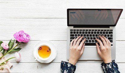 Person using laptop computer, pink roses on left side of table with white tea cup and saucer