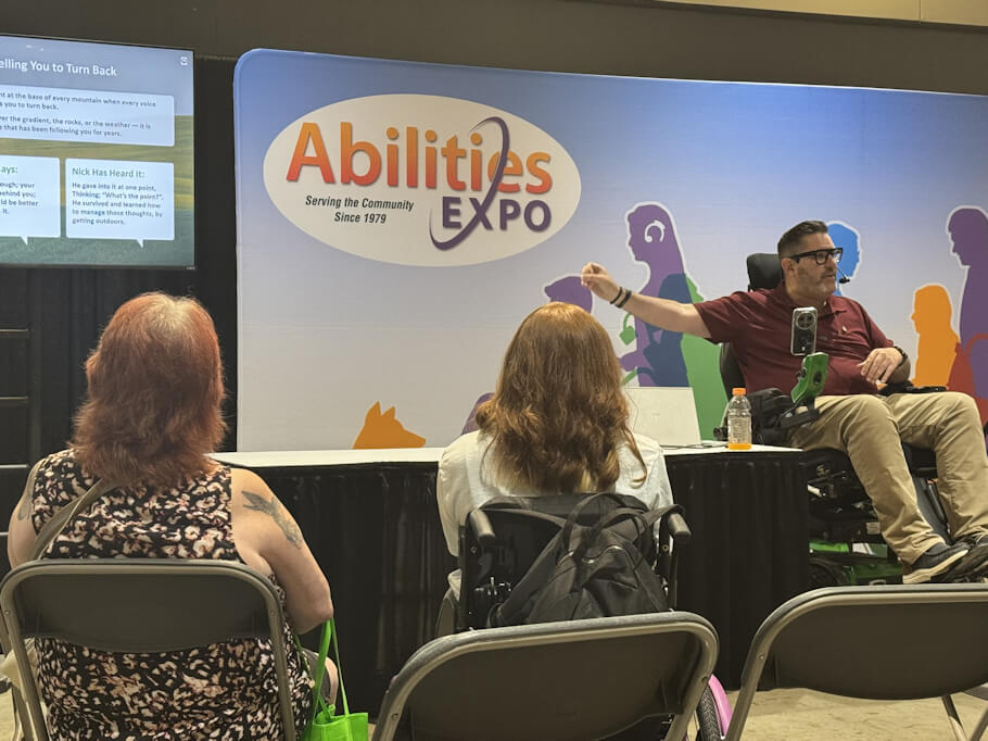 Nick Wilson, a Disabled Adventurer, using a power wheelchair sits at the front of a presentation space at Abilities Expo, a disability community event.