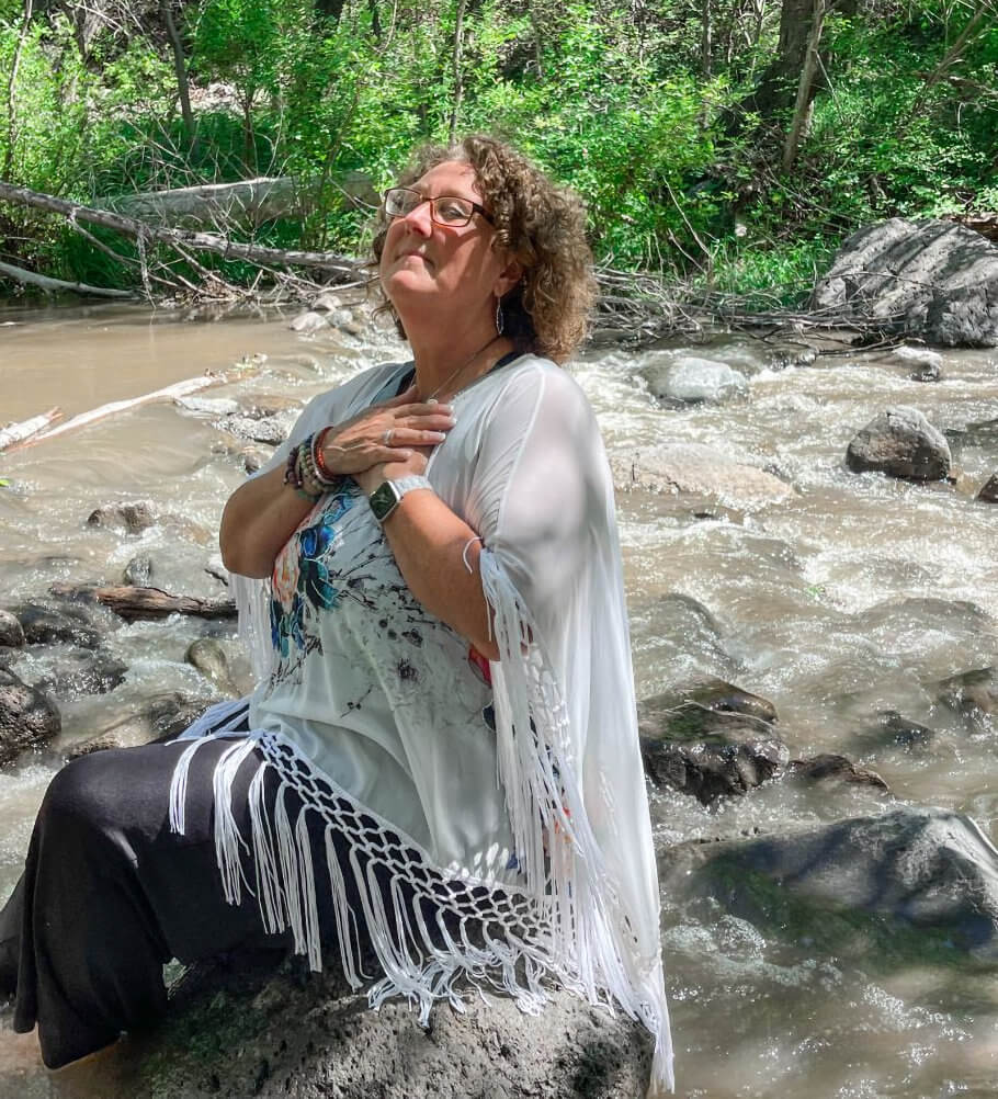 This image shows Dr. Teri Rouse sitting on a dark rock beside a flowing stream or river.