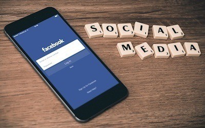 A cell phone with the Facebook app open next to Scrabble pieces arranged in the words Social Media.