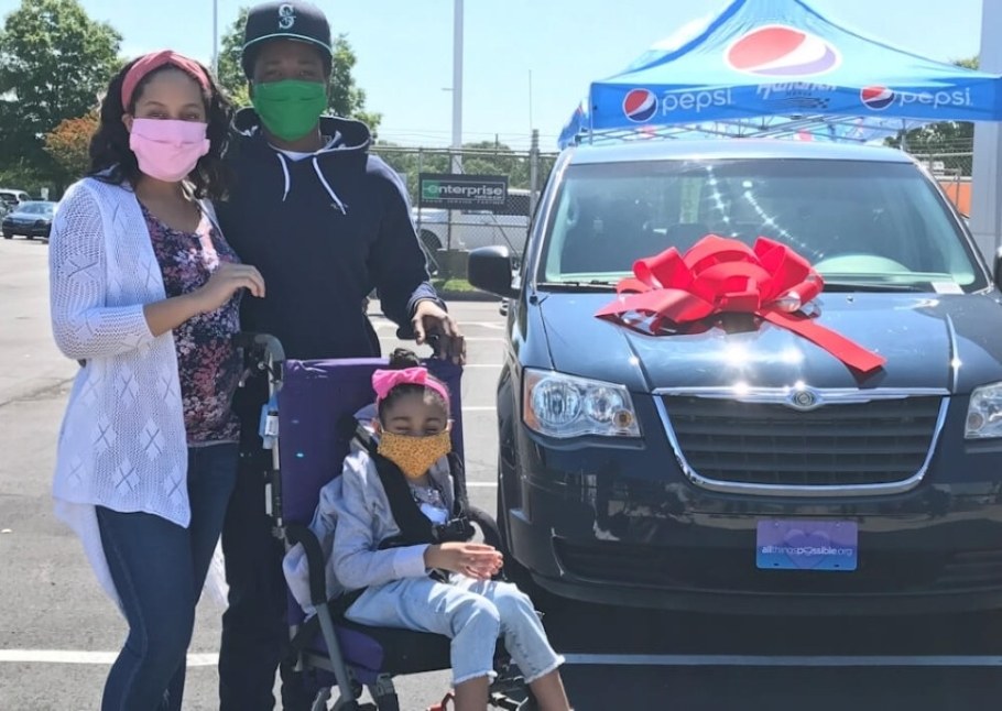 A family stands alongside their wheelchair-accessible van, with a red ribbon bow on the hood.