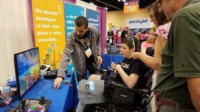 A man demonstrates video gaming to a teen boy in a wheelchair and his companions at a trade show.