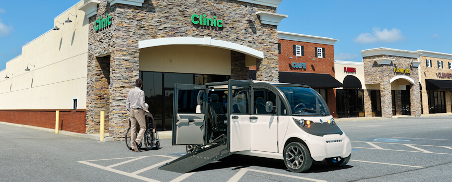This image shows a white GEM WAV (Wheelchair Accessible Vehicle) parked in front of a medical clinic in what appears to be a commercial strip center.