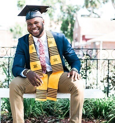 A smiling man in a graduation attire is sitting on a bench in a garden.