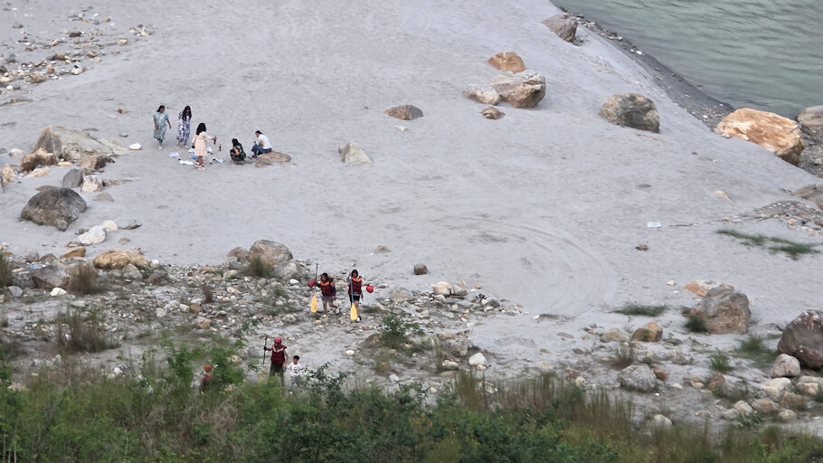 A wide, slightly elevated view shows a sandy and rocky riverbank beside calm, greenish-blue water on the right. Scattered large stones and patches of scrubby grass break up the pale ground.