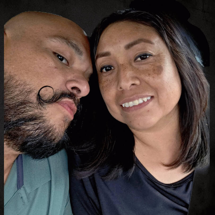 A close-up selfie shows two people sitting side by side inside a vehicle. On the left, a person with a short beard and a distinctive curled mustache leans their head affectionately toward the person next to them, looking in their direction.