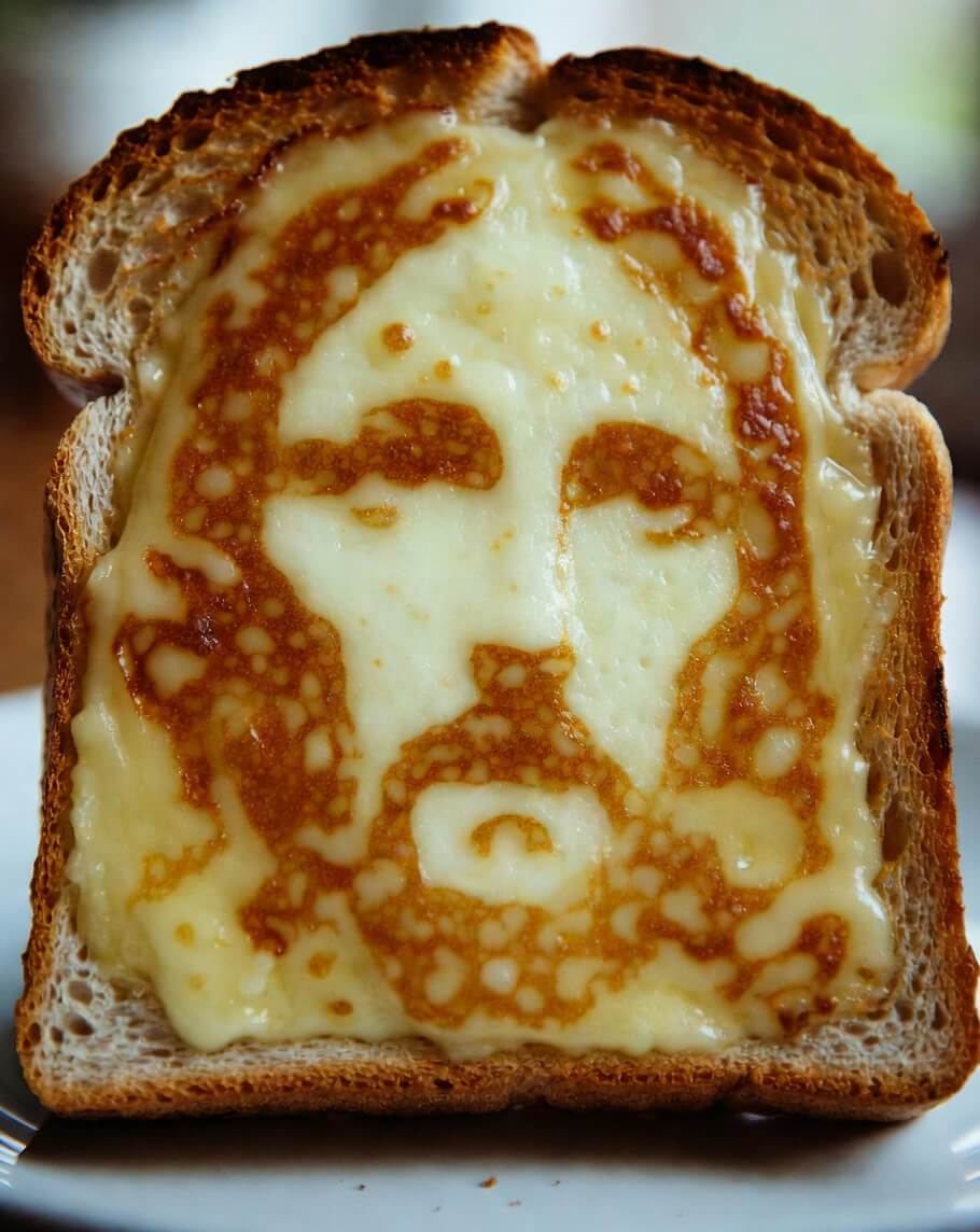Cheesus on Toast. This image shows a slice of toasted bread topped with melted cheese that has browned and bubbled during cooking, creating an unintentional pattern that resembles a human face - a phenomenon known as pareidolia.