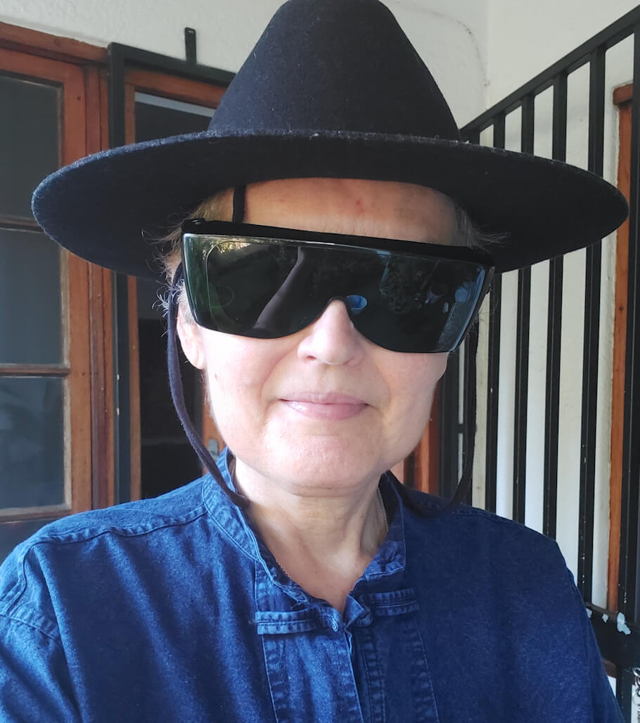 A close-up portrait shows artist and writer Joanne Bloch facing the camera with a gentle, relaxed smile. She is wearing a wide-brimmed black hat secured with a chin strap and oversized black wraparound sunglasses that cover her eyes and much of the upper half of her face.