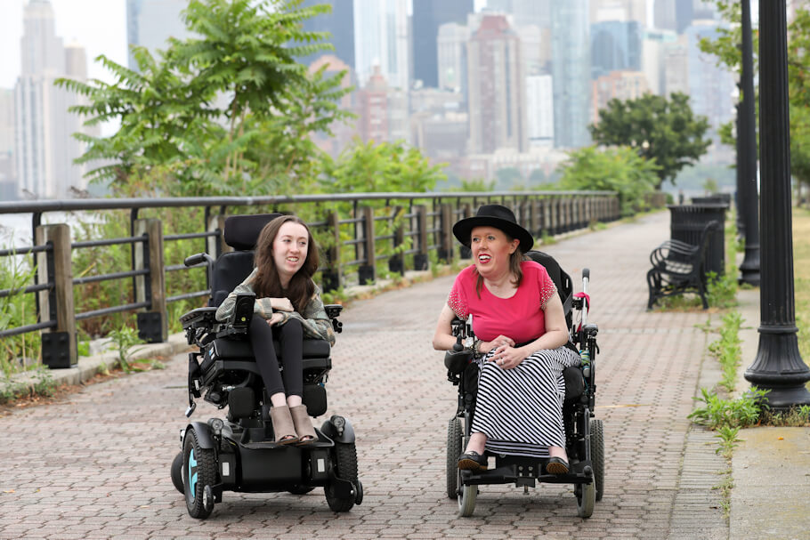 Avery Roberts and Kelly Berger , using powered wheelchairs, travel side by side along a wide brick waterfront promenade, smiling as they appear to enjoy a conversation.