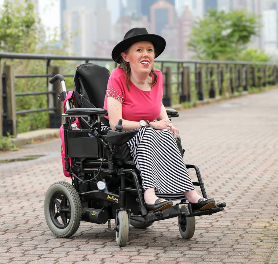 Kelly Berger sits in a black power wheelchair on a wide, paved brick walkway outdoors.