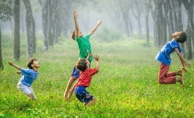 Four children in brightly colored clothes happily playing in a grassy wooded area.