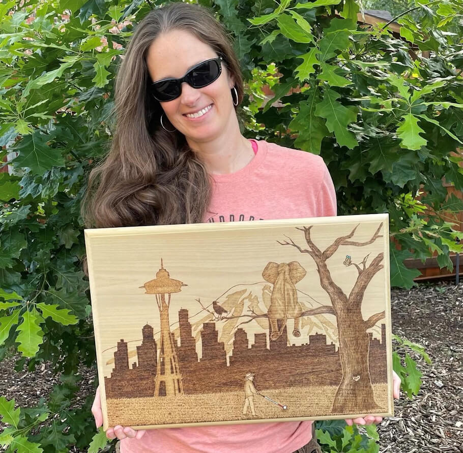 Krista Webb is standing outdoors in front of dense green leafy trees, holding a rectangular wooden artwork at chest height.