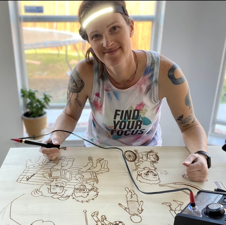 Pyrographer Krista Webb sits at a wooden table indoors, leaning slightly forward toward the camera while holding a pen-like tool connected by a cord to a small control box on the right side of the table.