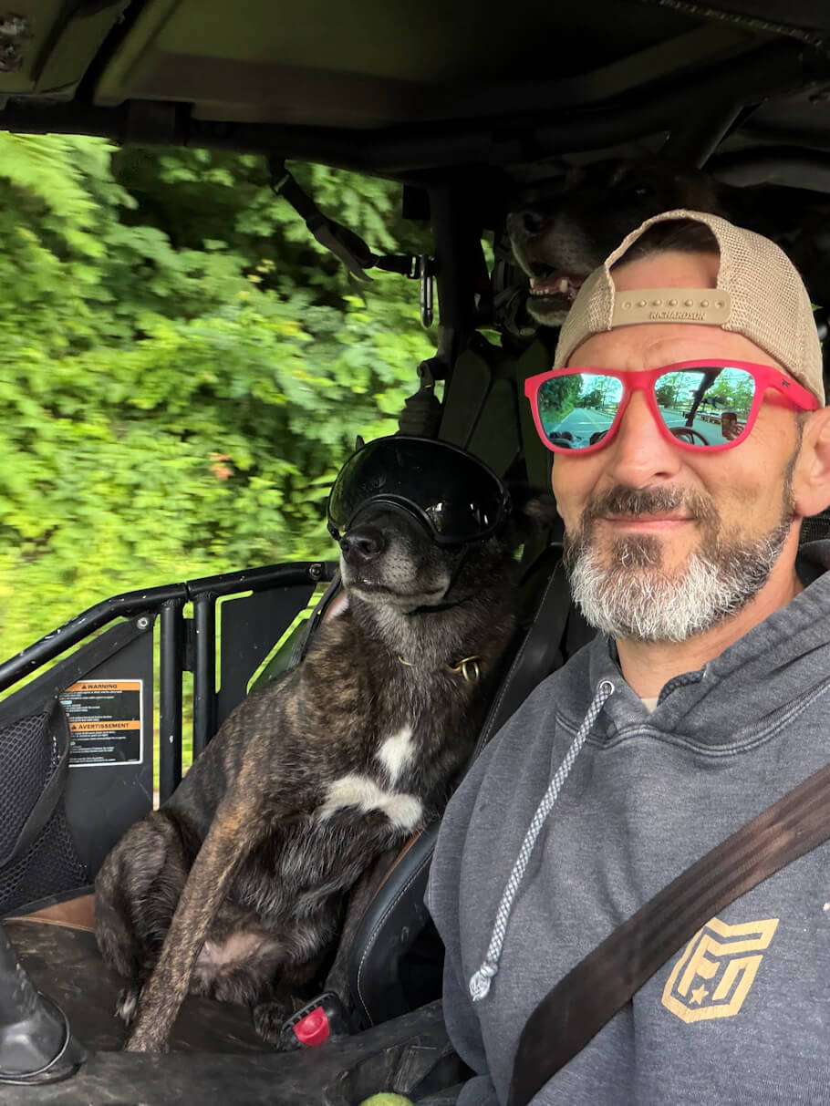 A smiling man wearing a gray hoodie, a backward tan baseball cap, bright pink mirrored sunglasses, and a seatbelt takes a selfie while riding in a side-by-side utility vehicle on a tree-lined road. Beside him, a brindle-colored dog with a white patch on its chest sits upright in the passenger seat wearing oversized black protective goggles, giving the scene a playful and adventurous feel.