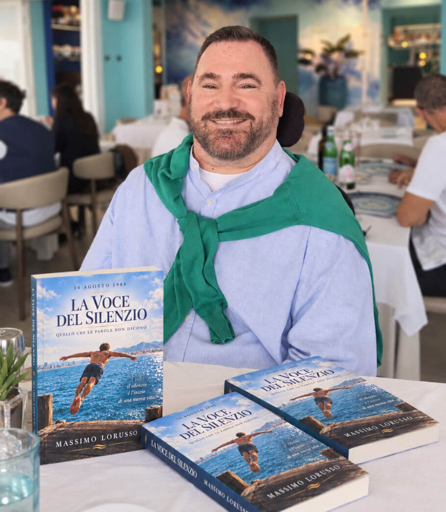 Massimo Lorusso sits at a restaurant table covered with a white tablecloth. He is wearing a light blue button-up shirt with a band collar over a white T-shirt, and a green sweater is draped over his shoulders with the sleeves loosely knotted across his chest.