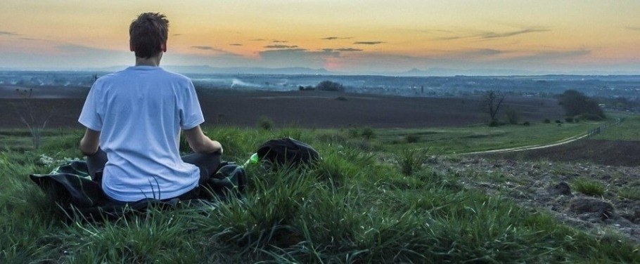 Man with his back to the camera meditating on a grassy hill overlooking a distant city.
