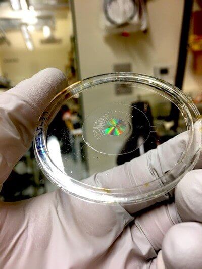 This is a photo of the metalens (made of silicon) mounted on a transparent, stretchy polymer film, without any electrodes. The colorful iridescence is produced by the large number of nanostructures within the metalens - Photo Credit: Harvard SEAS.