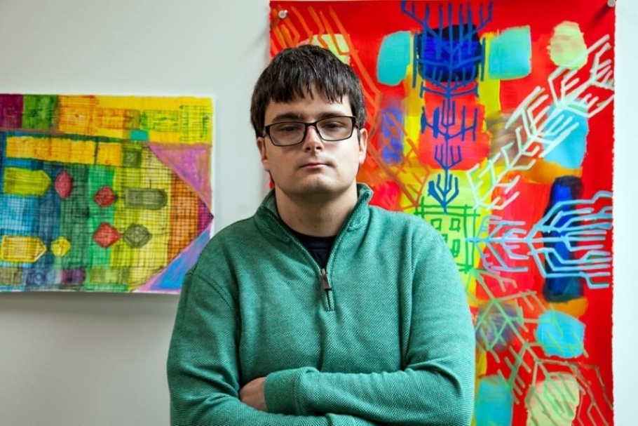 A photo of Michael Engebretson shows him standing in front of two of his pieces at Interact Gallery. He wears black-rimmed glasses and a turquoise pullover. He folds his arms and faces straight ahead. Sunlight pours into the space from the right, illuminating the vibrant colors of the works on the white wall behind him - Image courtesy of Bernie Wire, 2022.