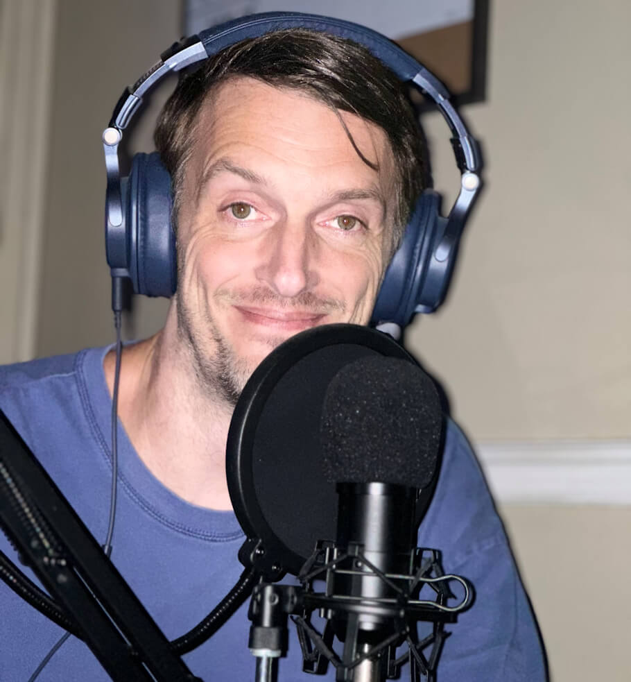 Michael Morris is seated at a microphone in a recording studio setting, wearing blue over-ear headphones and a blue t-shirt.