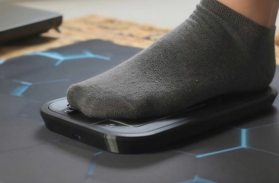 The image shows a close-up of a person's foot wearing a dark sock resting on a slim, rectangular assistive foot mouse placed on a desk mat or floor surface.