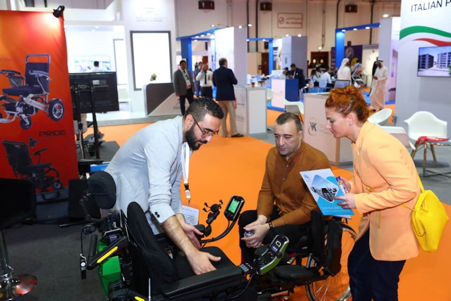 A person demonstrates an assistive device to a man in a wheelchair and a woman standing nearby.