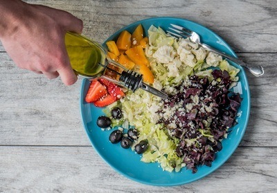 Person pouring oil from a bottle onto a plate containing vegetable and fruit salad.