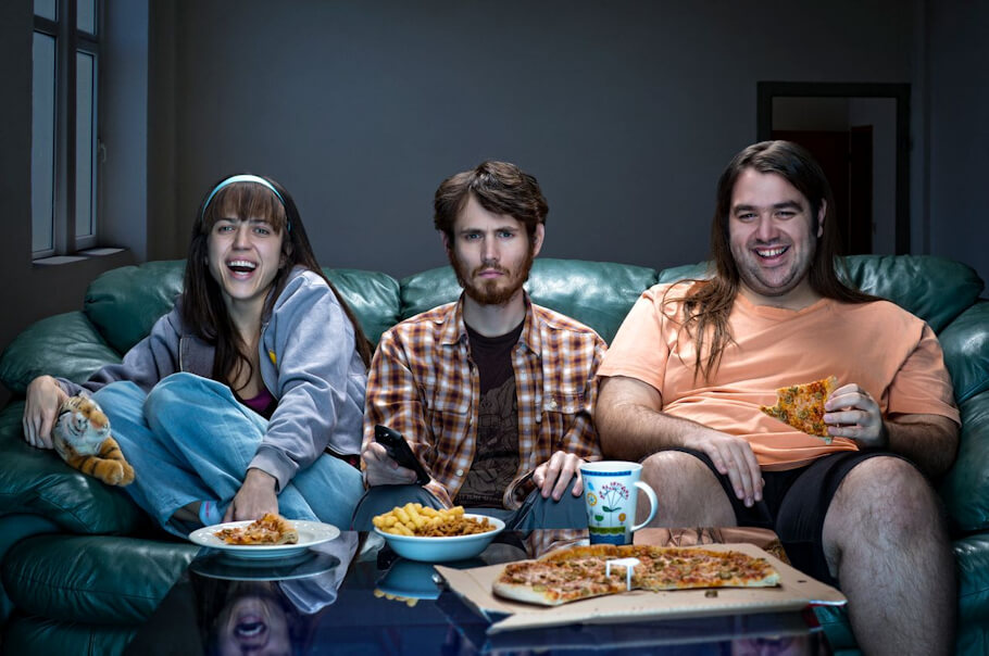In a dimly lit living room, three young adults are gathered on a plush, dark green leather sofa for a movie night.