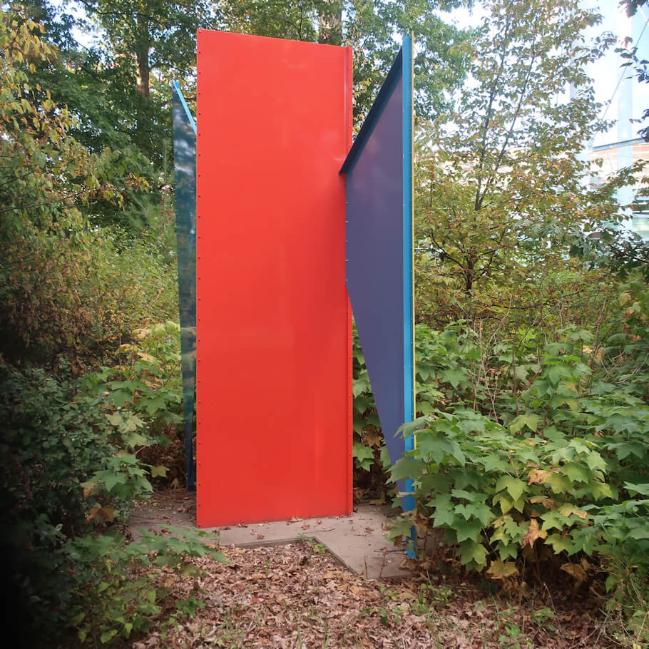 This photograph shows a modern abstract sculpture situated in a natural garden setting surrounded by trees and foliage.