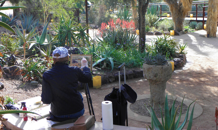 In this image, a person wearing a black quilted jacket and a checkered cap sits on a bench in a desert botanical garden, painting the vibrant landscape in front of them.