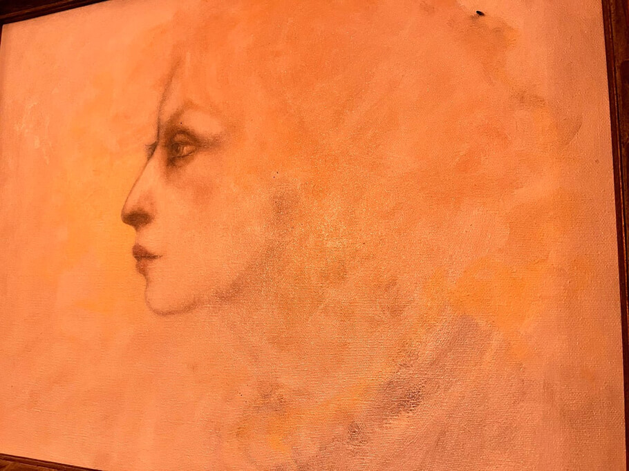 This image shows a framed painting on canvas shows a woman's profile facing left, rendered in soft, sketch-like detail against a warm, atmospheric background of peach, orange, and amber tones.
