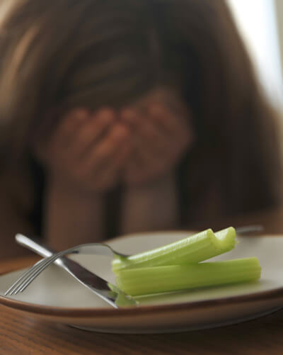 A plate with 2 small sticks of celery in front of a girl hiding her face in her hands.