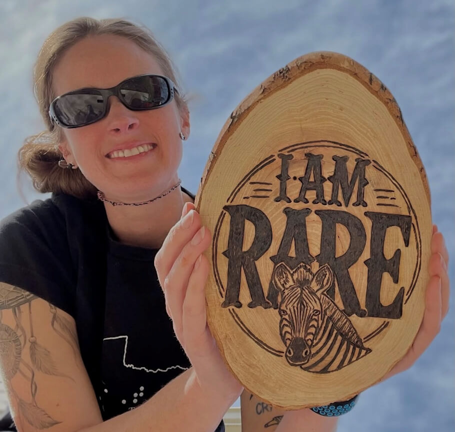 Pyrographer Krista Webb wearing dark sunglasses holds up an oval slice of natural wood with the bark still around the edges, presenting it toward the camera.