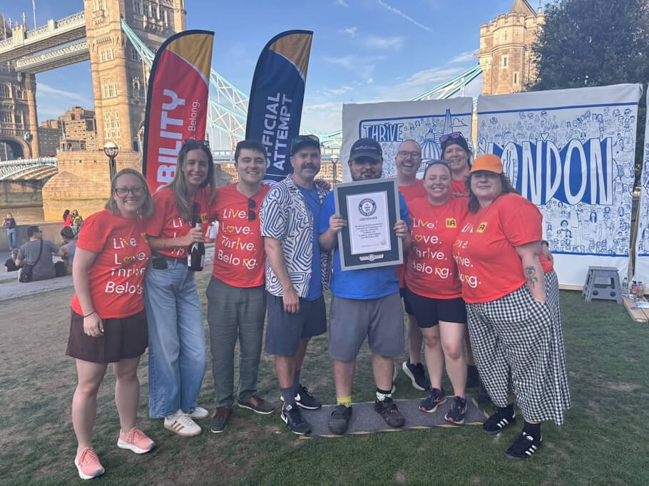 A group of nine people stands closely together on a grassy lawn beside the River Thames in London, with the distinctive twin towers and blue suspension cables of Tower Bridge rising in the background under a clear sky.