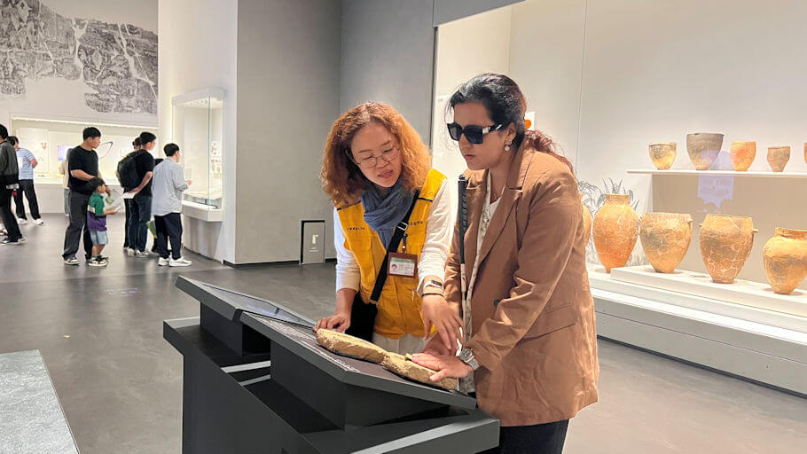 In this image, two women stand at a low black display podium inside a bright, modern museum gallery. The woman on the right is a visitor who is blind or has low vision, wearing dark sunglasses, a tan blazer and a white top, with a folding white cane slung at her shoulder; both of her hands rest on a pale tactile replica of an ancient stone tool set into the podium beside a panel of raised Braille text.