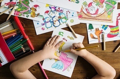 Image shows child coloring in a picture, colored pencils and completed colored pictures are scattered on the desk.