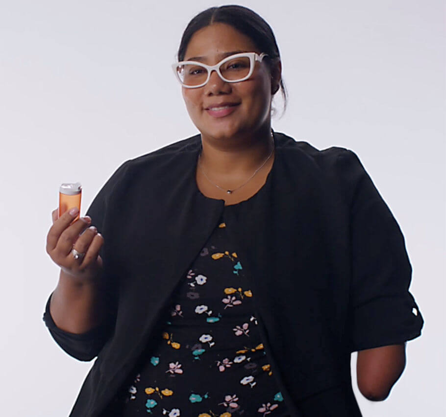 A smiling woman stands against a plain light gray background. She wears white cat-eye glasses, a thin silver necklace with a small pendant, and a black open jacket over a black top with a small floral print in yellow, pink, white, and teal.