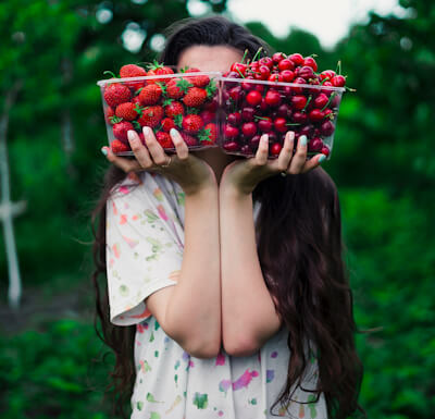 Young shy woman hiding her face behind two containers of strawberries and cherries.