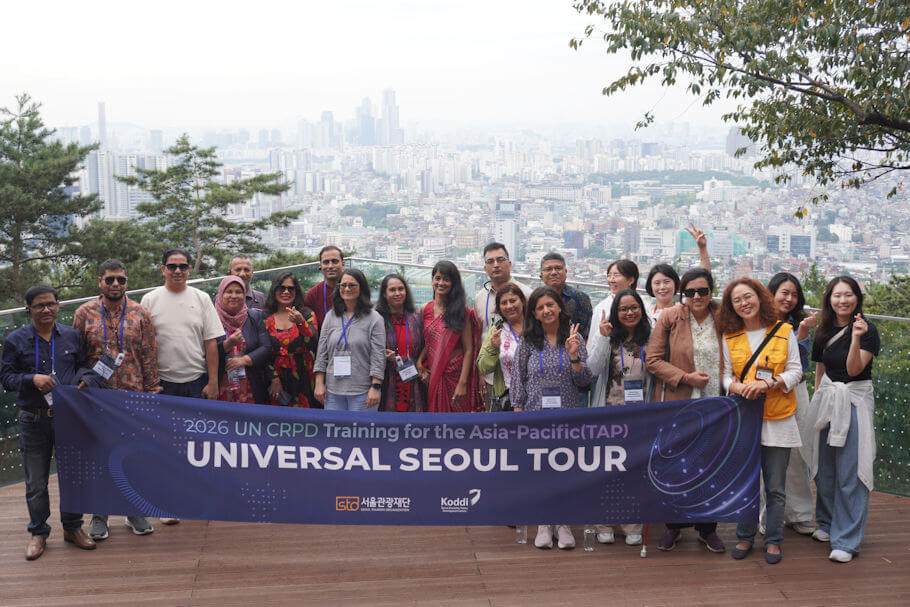 This photo is an outdoor group photograph on a wooden observation deck high above Seoul, with the city's dense rooftops and distant high-rise towers fading into a soft haze behind a glass railing and green pine and leafy branches framing the upper corners. About twenty people stand in two rows, dressed in a mix of business clothing and traditional South Asian and Korean styles, several wearing conference lanyards and dark sunglasses, some raising peace signs or finger hearts, with a white cane visible near the front and a staff member in a yellow volunteer vest at the right.