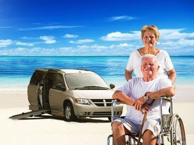 Woman standing behind man in wheelchair with van and beach in background.