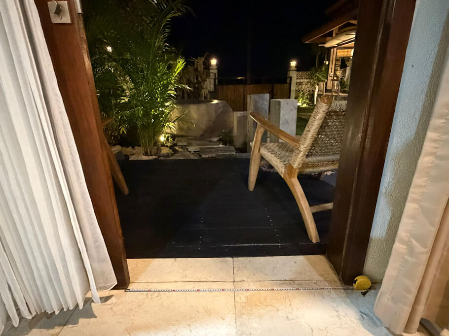 Viewed from inside the bedroom at night, wide double wooden doors open onto a dark timber deck that sits level with the pale stone interior floor, with a measuring tape stretched across the threshold showing the smooth step free transition.