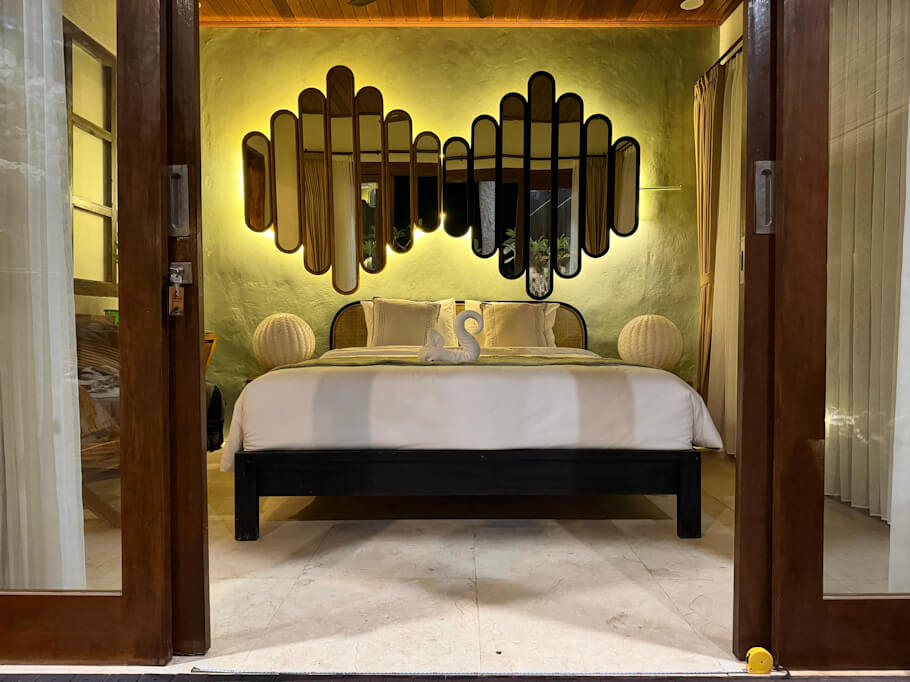 Accessible bedroom interior: Seen through open double wooden doors, a neatly made king size bed with crisp white linens, a rattan headboard, and a folded towel shaped like a swan sits centered against a softly lit olive green wall.